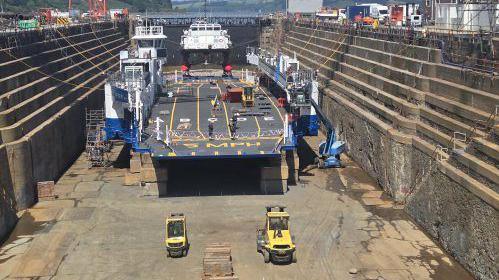 The Lynher in the dry dock. It is a car ferry and has five lanes labelled A,B,C,D,E. There are yellow construction vehicles on the ground. The vessel is blue and white. The walls of the dry dock are brown and have stairs on the right hand side. The ship is tied to the walls.