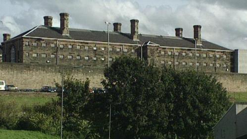 Brown prison block behind a large wall. The block has a lot of small windows and six chimneys.