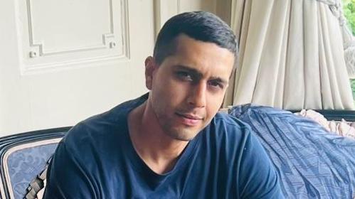 Kamran Aman wearing a blue top and sitting a sofa with a blue blanket behind him. He is looking at the camera and smiling.