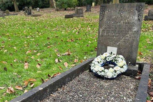 A grave that is inscribed 'in loving memory of Albert Willits, beloved husband of Dorothy Willits, who was shot in the execution of his duty', with further words below. A wreath of white flowers is based at the foot of the grave, which is surrounded by grass.