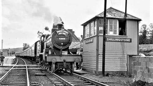 A black and white image from 1962. A 7F 2-8-0 S&DJR locomotive 53808 is on the track, with steam coming out of its chimney as it approaches a small hut, with a sign which reads "Shillingstone" on the front.