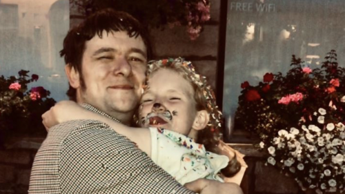 Rhys (left) and Millie and smiling and hugging tightly. Millie has face paint on and they're standing outside in front of flowers in window boxes at night.