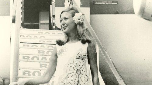 A woman walks down the stairs off an aeroplane. She wears the paper dress design from 1967 and has a large flower in her hair, looking off to the left of the photo. 