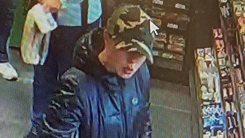 Ms Taylor wears a dark coat and camouflage cap. She stands in front of a counter which displays chocolate bars and sweets.