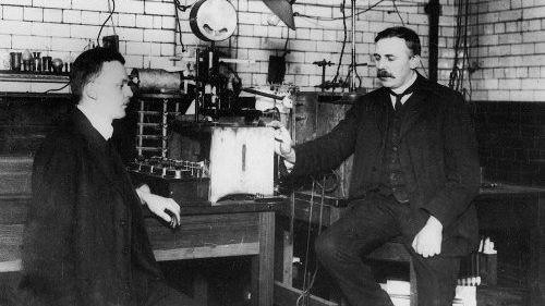 Ernest Rutherford (right) and Hans Geiger sit either side of a table of lab equipment in a black-and-white photo of a physics laboratory in Manchester in 1913.