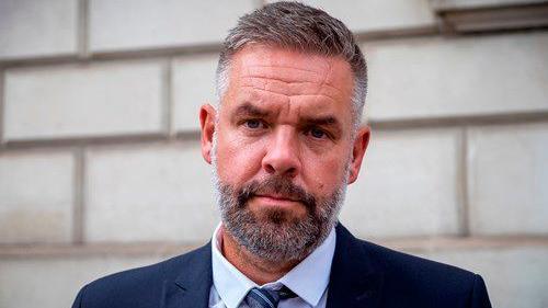 Lee Sims, the chair of the Humberside Police Federation, stands in front of a stone brick wall. He is wearing a dark blue suit, blue shirt and a grey and blue striped tie. He has short cropped, greying hair and a beard.