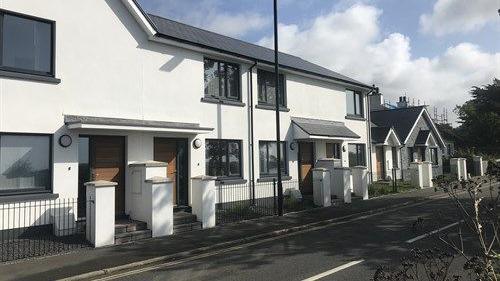 A row of white houses with fences and pillars at the front and steps leading to the front doors. They sit behind a pavement alongside a road, with a blue sky above them.