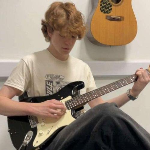 Wilf Fitchett sitting on a chair in a cream t shirt and black trousers. He is holding a cream and black guitar and is looking down at it as he plays. There is an acoustic guitar hanging on the wall behind him