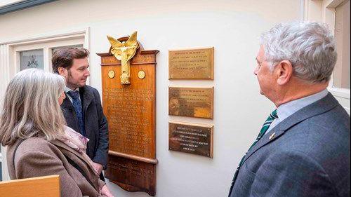 Two men and a woman stare at three golden plaques on a white wall, with a wooden board with names inscribed on in gold next to it.