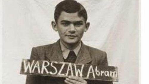 A black and white photo from 1945 showing Alec Ward holding up a chalk board bearing his birth name Abram Warszaw. He is wearing a collar shirt and a jacket and he has dark parted hair.