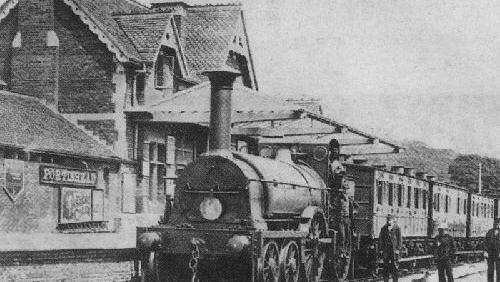 Huge locomotive steam train in front of an old railway station. It's an archive photo which is quite grainy and in black and white.