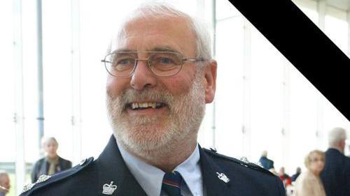 A man with a grey beard and short grey hair looking to left and smiling. He is wearing glasses and a blue jacket, blue shirt and dark green and red striped tie.