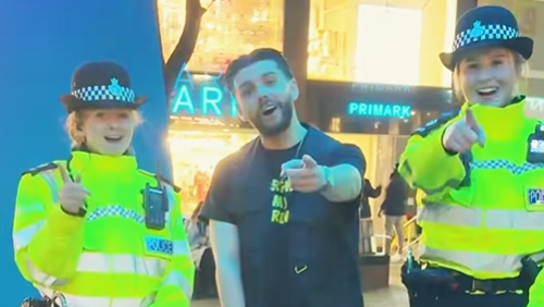 Leon Mallet wearing a black t-shirt and flanked by two female police officers in high visibility jackets. All are pointing to the camera and smiling.