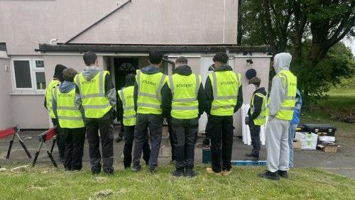 A row of students wearing hig-vis green vests stand outside a home where one of the new white fences is being installed.