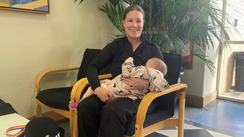 Sgt Kat Scrimshaw in a black shirt and black trousers. She is sitting in a chair. She is holding a small child in a pink onesie, and is breastfeeding. 