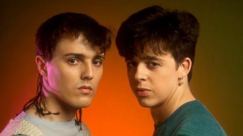Two men in 1980s style fashion, with haircuts typical of the time, face towards the camera, standing in front of an orange background.