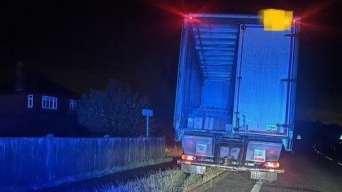 A lorry has come to a stop halfway up a grass verge beside a road at night. The lorry is lit blue by the lights of a police car, which is out of shot, and the red brake lights are on. The back doors are half open and boxes can be seen inside. To the left is a fence and a house can be seen in the distance.