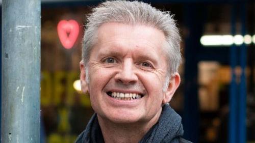 A man looks at the camera and smiles with his teeth. He is wearing a grey scarf as he stands outside.