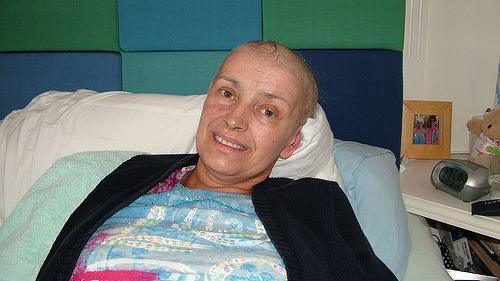 Olwen Kramer lies in a hospital bed. She has lost her hair because of cancer treatment. She is wearing a black cardigan with a white, blue and red patterned shirt underneath.  