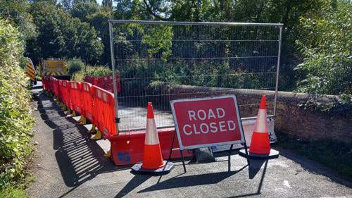 A bridge is closed off from traffic with temporary gates. In the front of the picture is a red sign saying Road Closed.