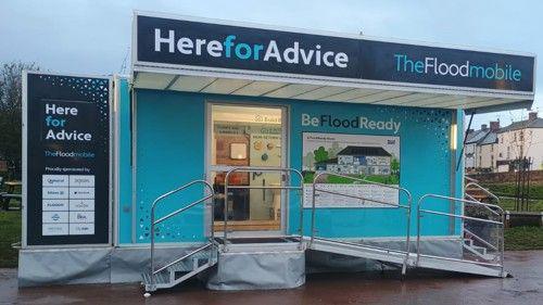 An exhibition trailer, with blue and white branding which identifies it as a 'Floodmoible'. There are information boards outside and inside the trailer and there are houses in the distance behind it.