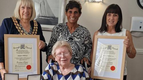Four woman, one seated, alongside each other in a room with a picture of a sailing ship on the wall. Fatima Whitbread stands behind her mother Margaret who is in a wheelchair. 