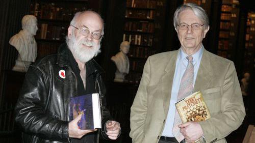 Sir Terry is dressed in a black leath jacket next to Colin Smythe who is wearing a tan coloured blazer and blue shirt. They are both holding hardback copies of his book and are stood together in a library