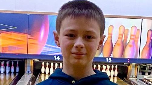 A close-up photo of a boy looking straight at the camera. He has short brown hair and he is wearing a blue hoodie. He is in a ten-pin bowling alley, with lanes and sets of pins visible behind him.