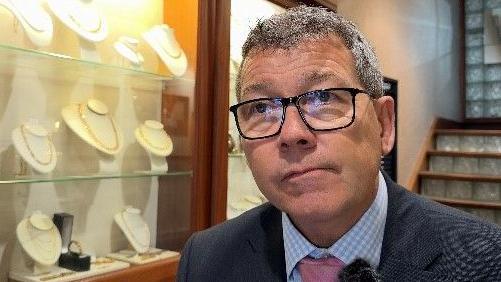 Jeweller Tim Hopper stands in his shop next to a glass cabinet, in which a range of gold necklaces are on display. He has short hair and wears dark-rimmed glasses, a dark-grey suit, a blue-checked shirt and a pink tie.