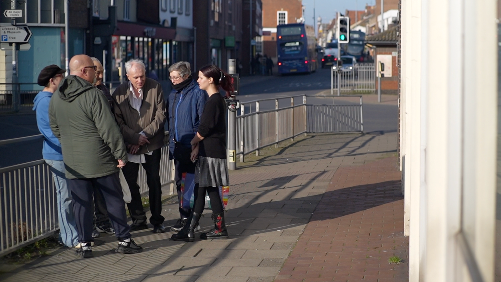 A group of people stand outside the library in a high street. The pavement and the road are separated by railings and the group stands next to the railings as a bus goes by in the distance