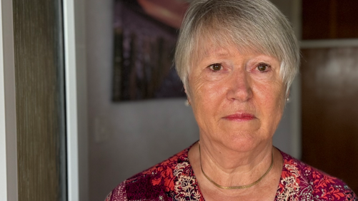 The head and shoulders of Sheila Jane. She is standing in her doorway and looking directly into the camera. She is wearing a red floral top or dress.