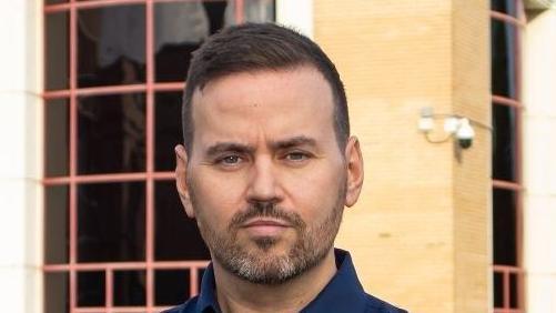 Gavin Callaghan smiles at the camera. He has short dark hair with a beard and wears a navy blue shirt. A yellow brick building can be seen behind him with large windows.