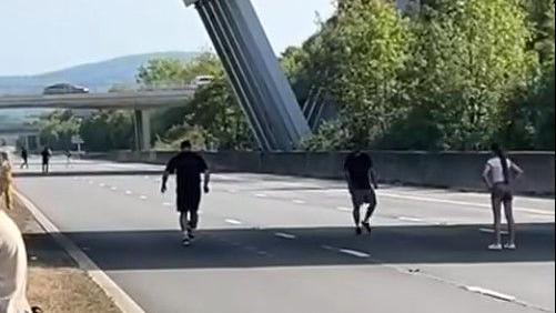 Three people stand close to each other on a closed road, with one person kicking a ball with their back to the camera. Another three people on the road can be seen in the background.