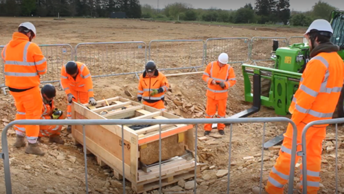 A stone coffin in a wooden frame next to a forklift truck with archaeologists in hi-vis orange jackets and trousers surrounding it