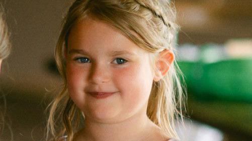 A head and shoulders picture of a seven-year-old girl. She has shoulder-length blonde hair and is smiling to the camera.