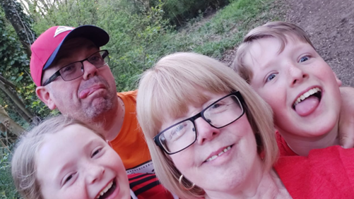 A mum and dad with their son and daughter pull silly faces at the camera as they take a selfie in the woods