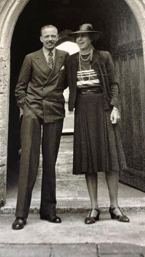 A black and white photo shows a man in a suit and a woman wearing a hat and pearls standing in front of the stone archway at Dartington Hall. They are both smiling.