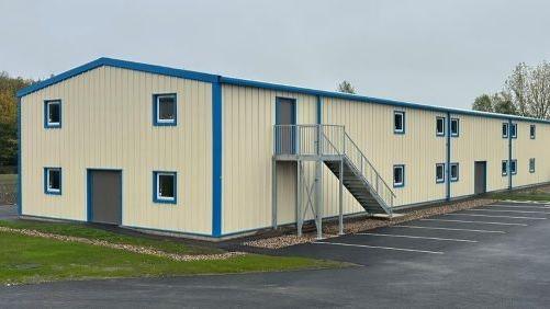 A cream, warehouse-style building with blue-painted window and door frames. An external stairway leads up to the first floor, with car parking immediately below the stairs and a patch of lawn to the left.