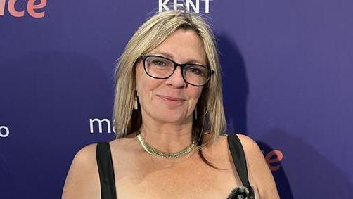 Jo Hogbin has blonde hair and is wearing a black dress and black acetate rimmed spectacles