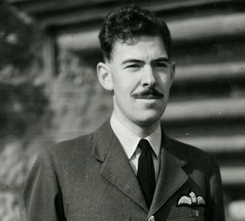 A black and white picture of John Cruickshank in his RAF uniform, staring just off camera. He has a dark moustache and dark hair. He is only visible from the neck up.