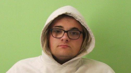A police picture of Jessica Garnham-Burton. She is wearing a white hooded jumper with the hood up. She has blonde hair and black glasses.