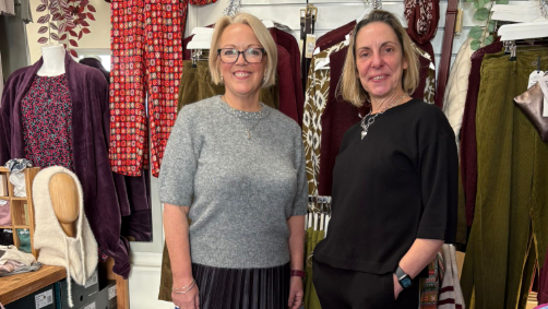 Emma standing with a second woman in the middle of rails of clothes in her shop, both are smiling to camera