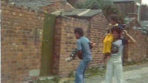 Two people in jeans and t-shirts. One carrying a child in their arms, another carrying a child on their shoulder. The group stands in a small road behind brick houses.