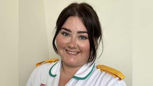 Kellie-Ann is in her white nursing uniform with yellow pads on her shoulders. She has dark brown hair which is tied back, dark eyebrows and blue eyes.