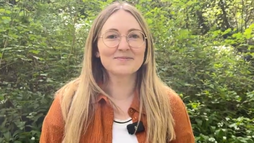 Becca is standing in front of a green hedge in the sun. She is wearing an orange cardigan and has long, blonde hair and glasses.