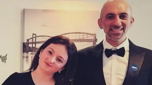 The image shows two people dressed formally, standing indoors. One person is wearing a black suit with a white shirt and a black bow tie, and there is a small blue badge pinned to the lapel. The other person is wearing a black dress.