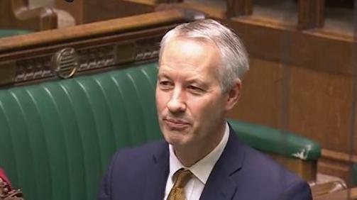 Gideon Amos MP for Taunton and Wellington addressing Parliament in a navy suit and a gold tie 