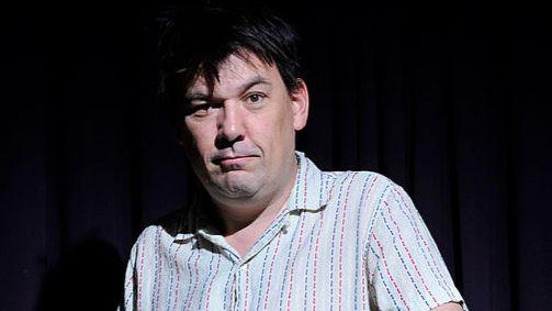 Graham Linehan wearing a patterned shirt standing in front of a dark background