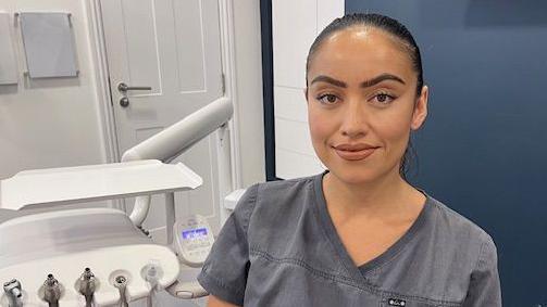 Kath Stahl sits in her dental surgery. Her black hair is tied back and she wears grey scrubs. Dental equipment can be seen to her left. 