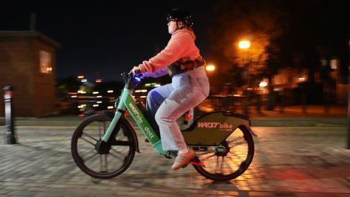 A woman in blue jeans, pink sweatshirts and black helmet rides a Dott and West Bike branded e-bike along a cobbled road at night time.  
